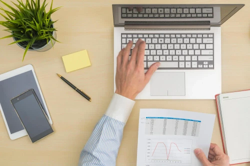 Pessoa teclando no notebook olhando um gráfico impresso